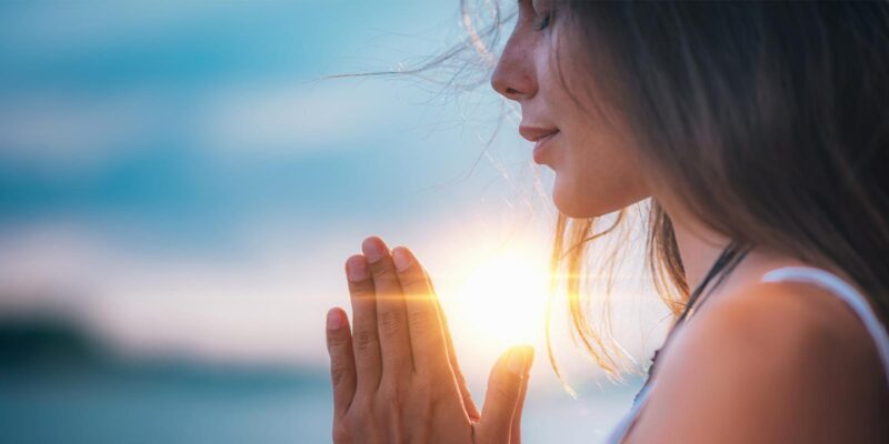 woman outside by the ocean with her hands together in a meditative state practicing mindfulness
