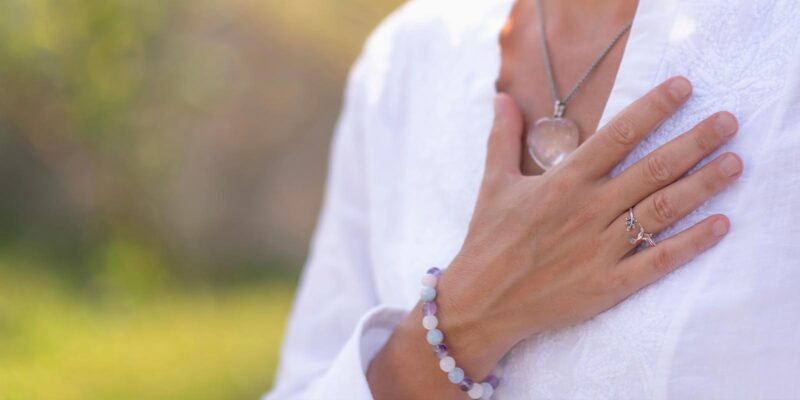 Woman wearing a white shirt and crystal necklace outside holds her hand over her heart to show gratitude for the things in her life.