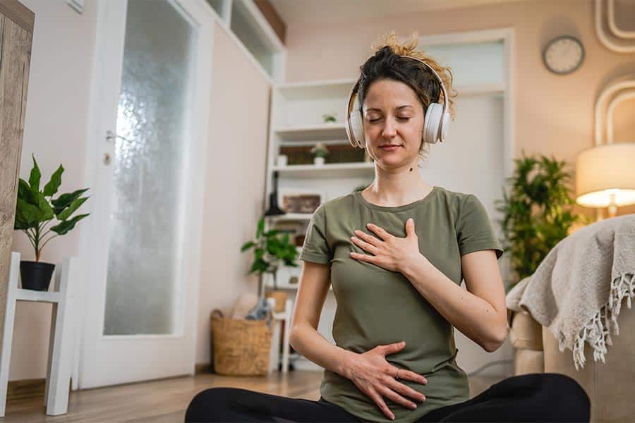 Woman practicing diaphragmatic breathing in her living room. She is listening to a mindfulness meditation with her headphones.