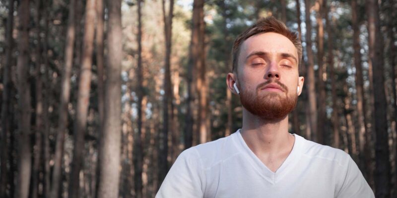Man with a beard sitting outside in a wooded area practicing mindful breathing with his eyes closed.