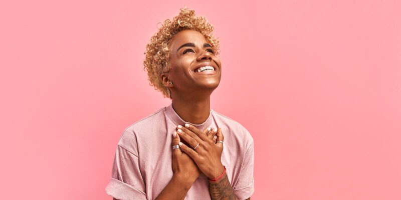 A person with curly blonde hair on a pick background clutches her chest and smiles to show gratitude for their life.