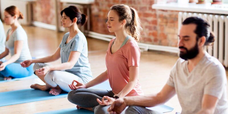 Coed group of four people practicing yoga, sitting in the lotus position. This image shows four people using yoga to benefit their addiction recovery plans.