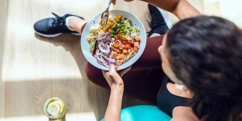An aerial view of a brunette woman in workout clothes, enjoying a colorful and nutritious salad, symbolizing the importance of healthy eating in recovery and overall wellbeing.