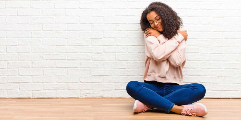 Image of a woman sitting on the floor against a white brick wall, embracing herself in a comforting hug, symbolizing self-compassion and emotional support.
